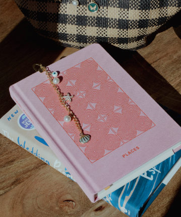 Pink book with a gold charm bookmark featuring shell, pearl, and colorful accents, next to a checkered bag and other books.