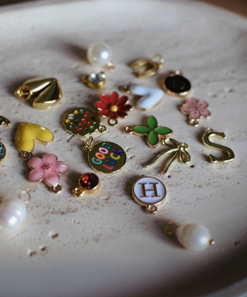 Collection of colorful and personalized charms featuring flowers, hearts, 'Good Luck' designs, initials, and pearl accents in gold settings.