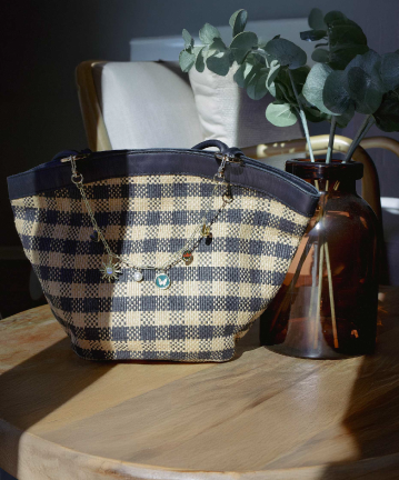 Stylish woven checkered handbag with a charm bracelet featuring floral, heart, and colorful charms, placed on a wooden table with a vase of greenery.